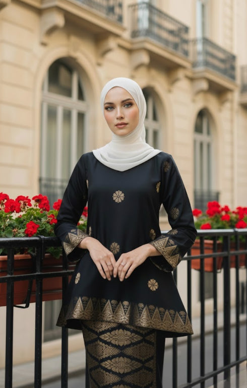 Woman wearing a Kurung Kedah Black Gold showing songket woven design and fit in front of a building with red flowers.