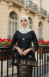 Woman wearing a Kurung Kedah Black Gold showing songket woven design and fit in front of a building with red flowers.