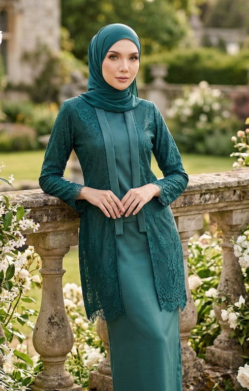 Woman styling kebaya Kutu Baru Teal Green front view with long straight sleeve and shoulder pad detail standing in a garden with flowers and stone architecture.