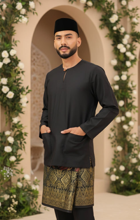 Man wearing a Baju Melayu Teluk Belanga traditional outfit Bllack with a Sampn 3D, standing in front of floral decorations.