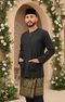 Man wearing a Baju Melayu Teluk Belanga traditional outfit Bllack with a Sampn 3D, standing in front of floral decorations.