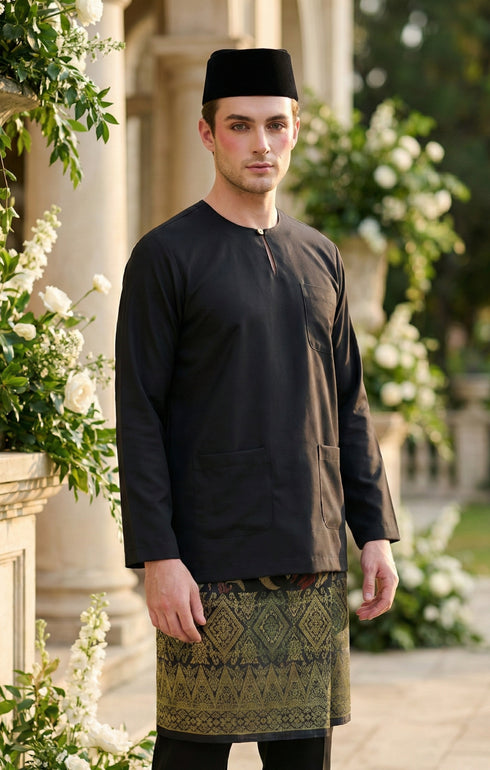 Man wearing a black Baju Melayu Teluk Belanga long-sleeve standing outdoors with greenery and flowers in the background.