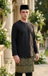 Man wearing a black Baju Melayu Teluk Belanga long-sleeve standing outdoors with greenery and flowers in the background.