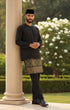 Man in traditional attire Black Baju Melayu Teluk Belanga standing next to a column with greenery in the background