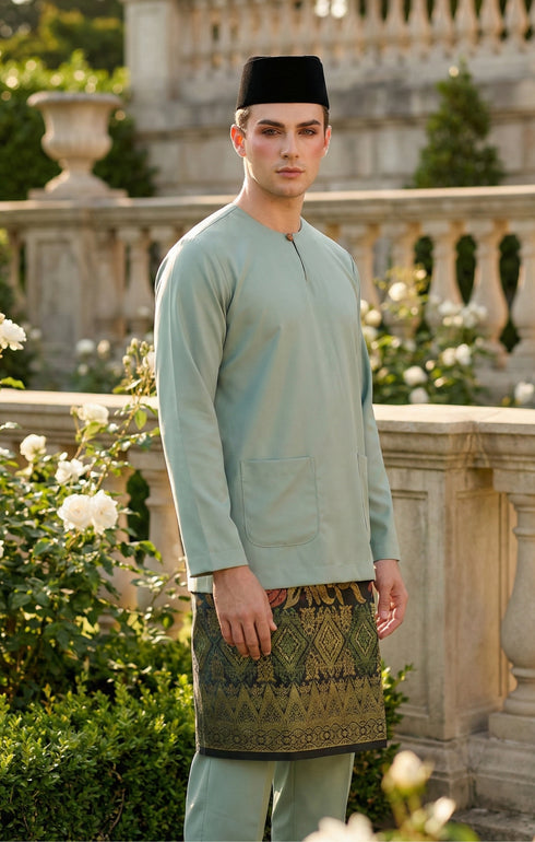 Man wearing a traditional outfit Baju Melayu with a Dusty green top and sampin and songkok in an outdoor setting.