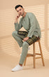 Man sitting on a wooden stool wearing a Dusty green Baju melayu Teluk Belanga outfit with with Malaysian Tradional Sampin in a beige background