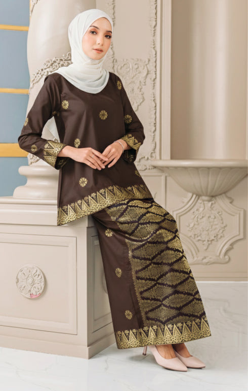 Woman wearing a Kurung Kedah Dark Rich Brown Gold traditional outfit with a hijab, standing against an elegant interior background.
