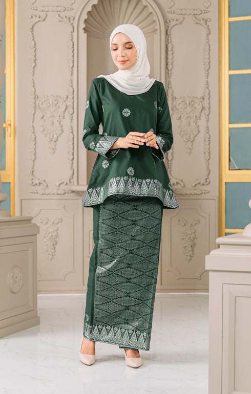 Woman wearing a Kurung Kedah Dark Emerald Green Silver traditional outfit with a matching Shawl Hijab in an elegant interior setting.