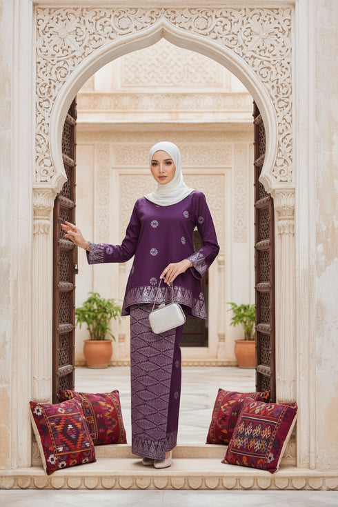 Woman in a Kurung Kedah Magenta Silver outfit standing in an ornate archway with decorative cushions.