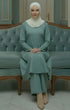 Woman Kebarung Klasik Plain Sage Green outfit sitting on a light blue couch in a decorated room.