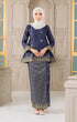 Woman wearing a Kurung Kedah Blue Black Gold highlighting long straight sleeve and silhouette traditional outfit with a matching pearl white instanl shawl scarf in an elegant interior setting.