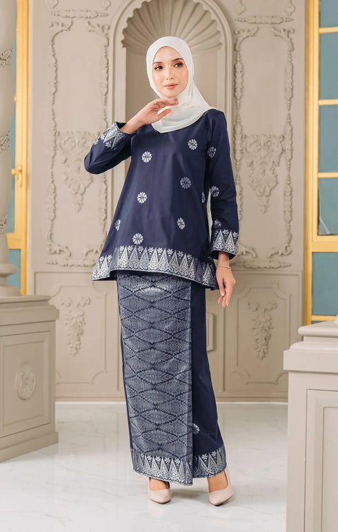 Woman wearing a Kurung Kedah Blue Black Silver traditional outfit with a matching pearl white instanl shawl scarf in an elegant interior setting