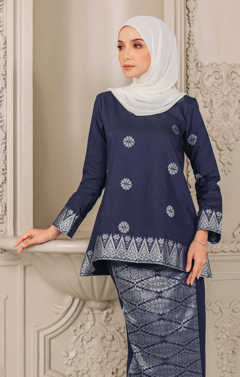 Woman wearing a Kurung Kedah Blue Black Silver traditional outfit showing fabric close up and texture with pearl white instant shawl in an elegant interior setting.