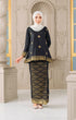 Woman wearing a Kurung Kedah Black Gold traditional outfit with a pearl white shawl hijab in an elegant interior setting.