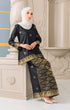 Woman wearing a Kurung Kedah Black Gold showing songket traditional outfit with a shwal hijab, standing against an elegant interior background.