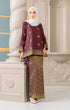 Woman in Traditional Kurung Kedah Dark Maroon Gold highlighting long straight sleeve and silhouette outfit with instant hijab standing against an ornate wall.