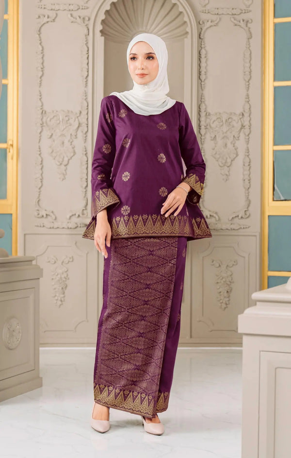 Woman wearing a Kurung Kedah Magenta Gold traditional outfit with gold accents against an ornate wall.
