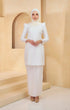 A woman wearing a Pearl White, solid-patterned traditional kurung moden with a round neckline and long sleeves, standing in a modern setting.